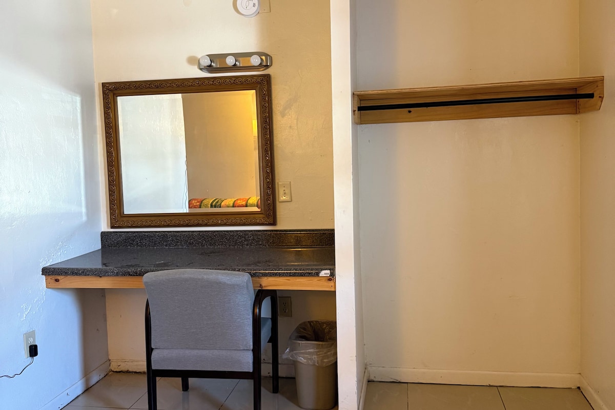 A small, compact room with a kitchenette area, a chair, a trash bin, and a mirror on the wall.