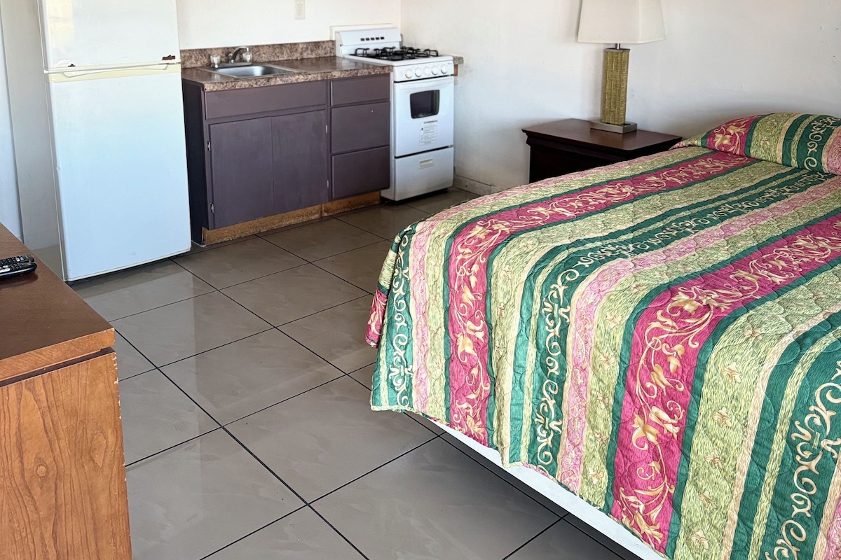 A compact hotel room with a kitchenette, a bed with a colorful bedspread, a nightstand with a lamp, and a wooden dresser.