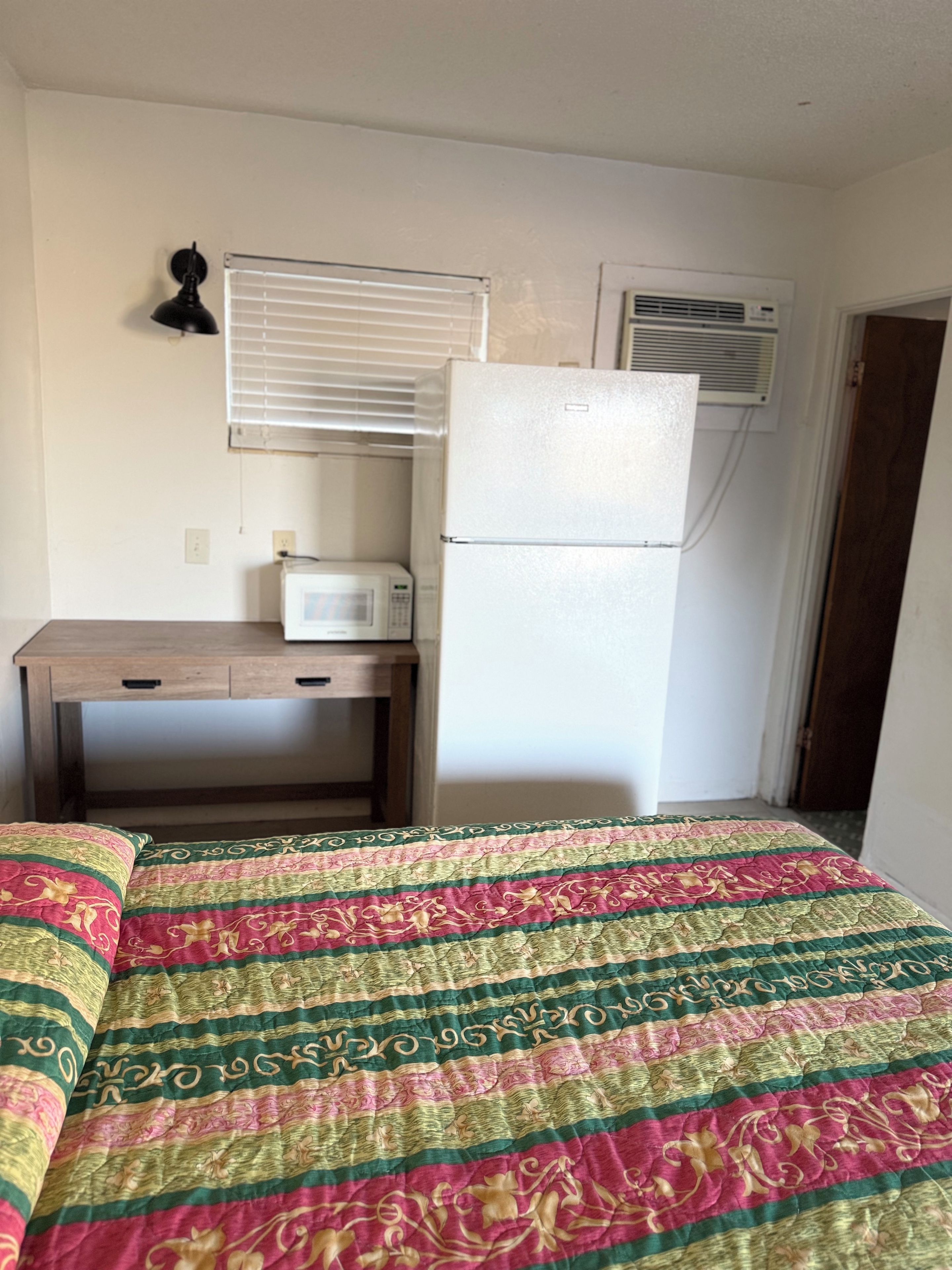 Una habitación con cama, escritorio, refrigerador, microondas y ventana con persianas.
