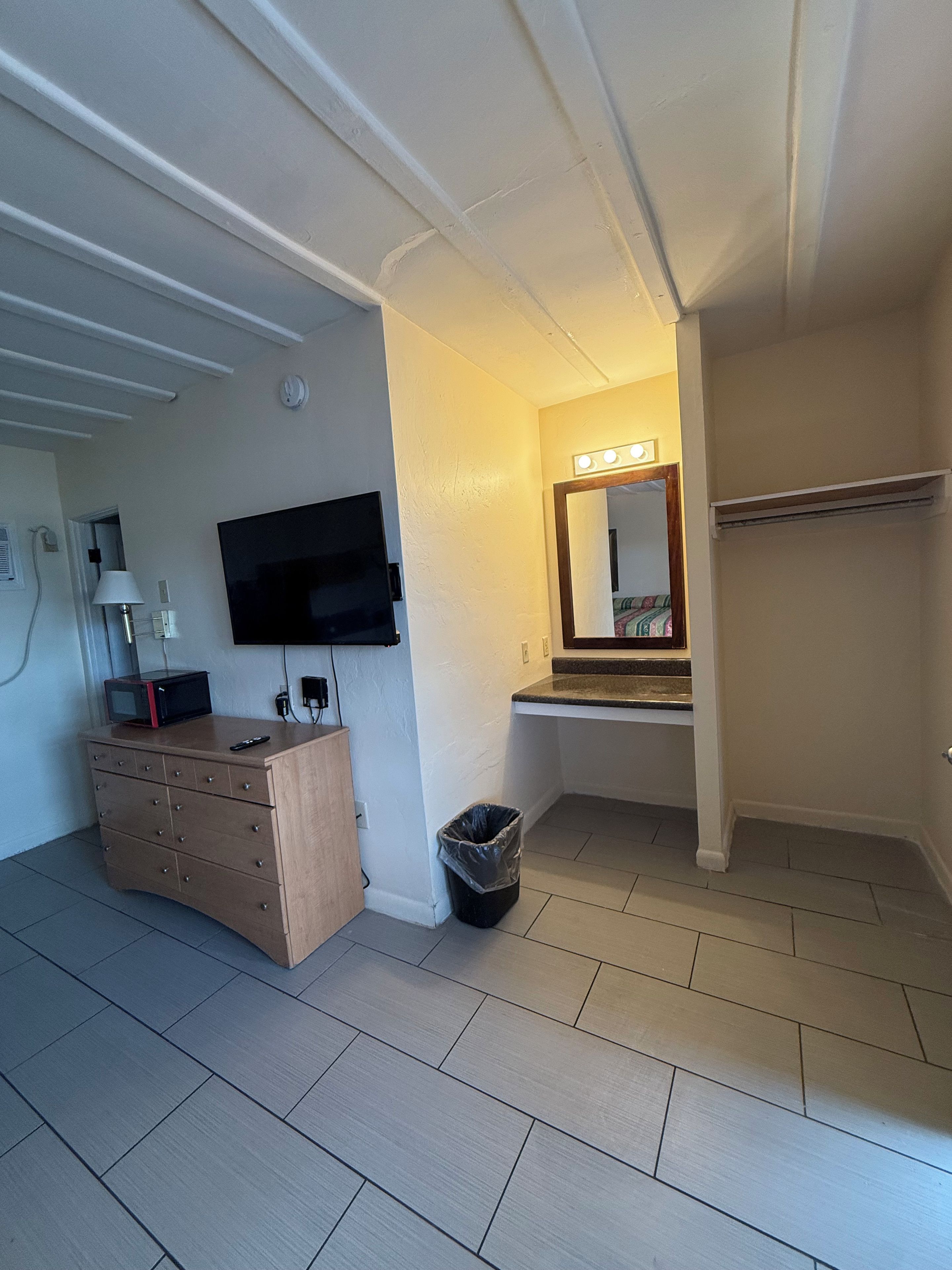 A hotel room with a flat-screen TV mounted on the wall, a wooden dresser, a trash bin, and a vanity with a mirror and lights.