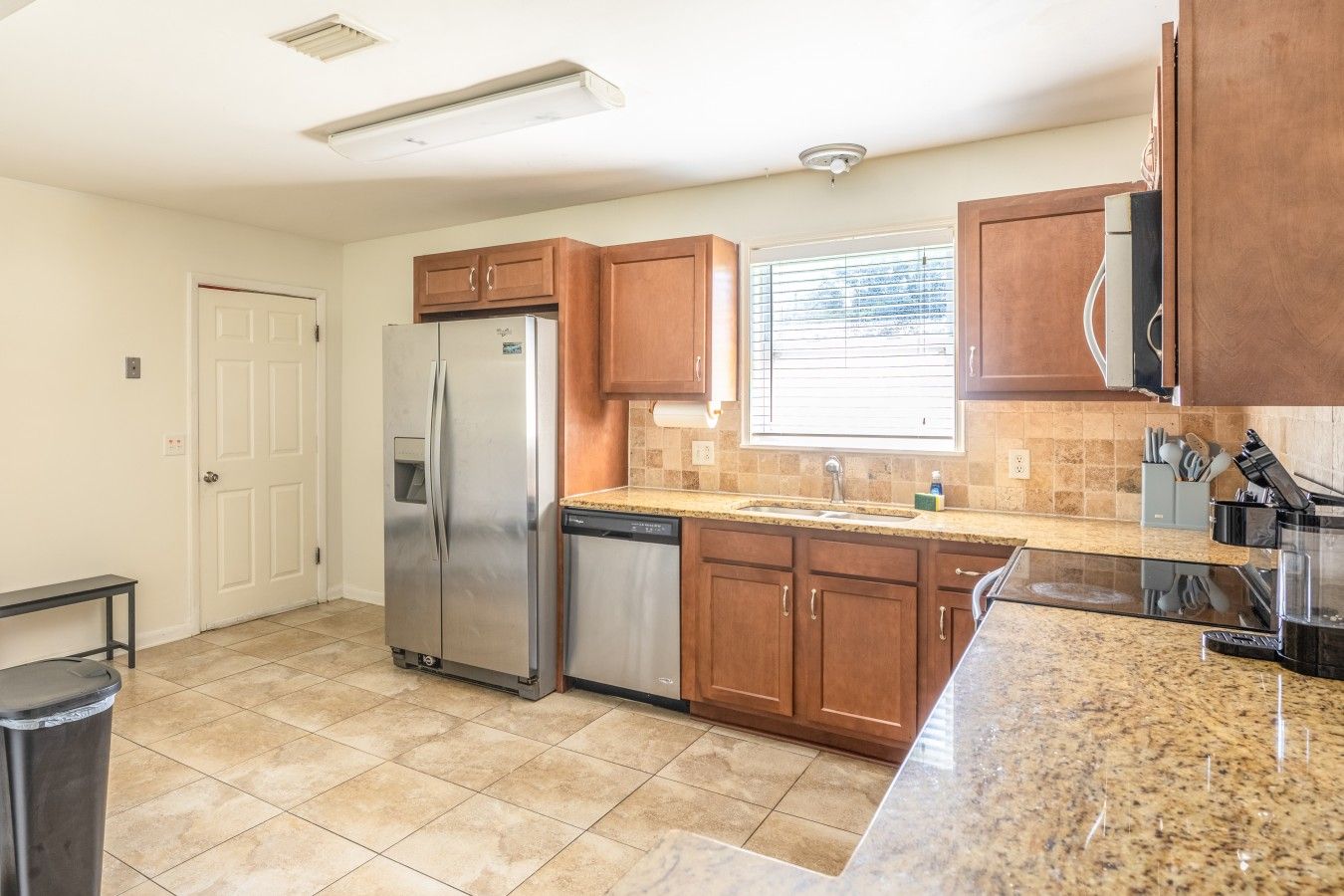 A kitchen with a stainless steel refrigerator, a dishwasher, wooden cabinets, a granite countertop, and a window with blinds.