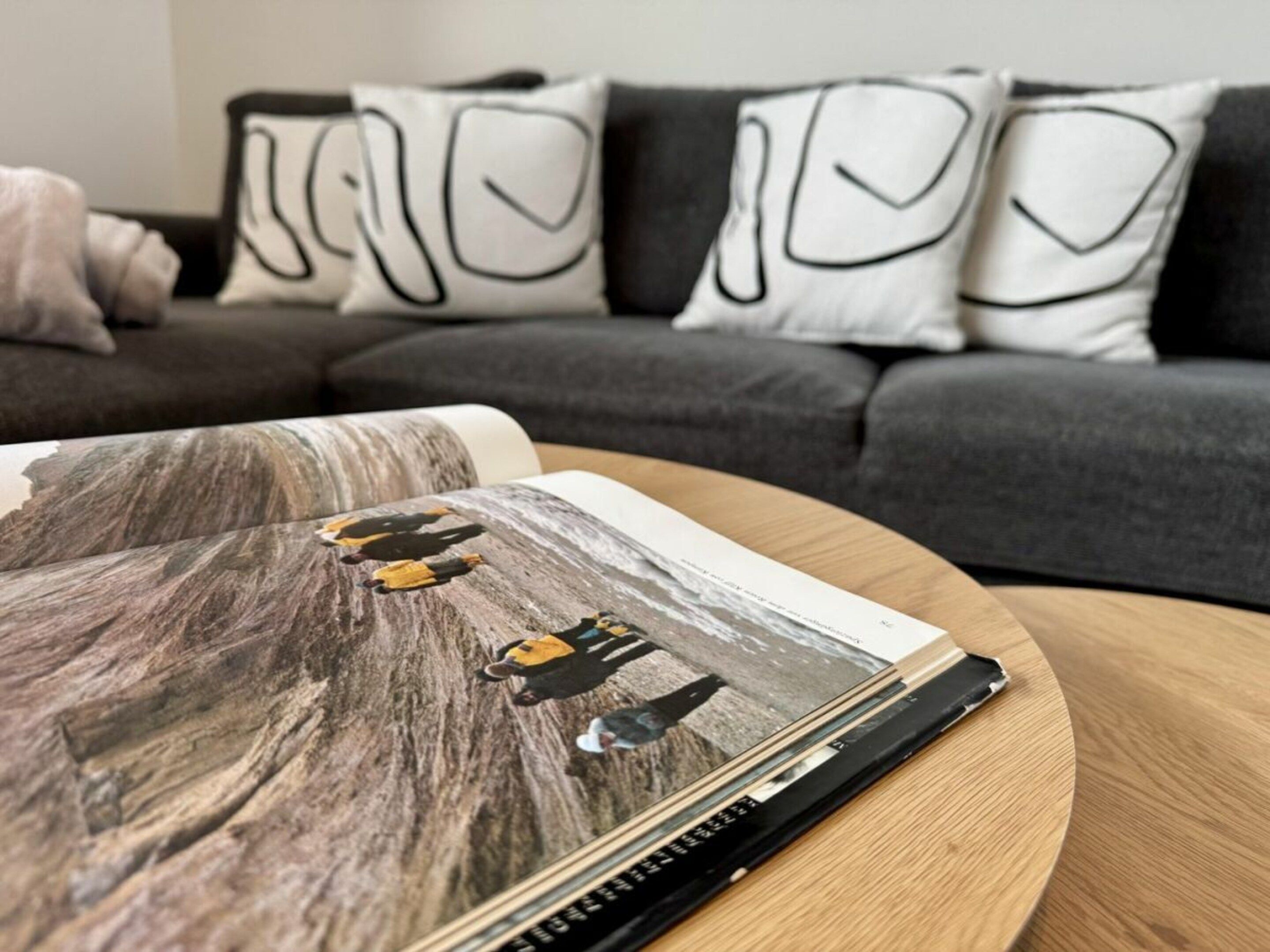 Ein Buch mit Holzmuster-Einband auf einem runden Holztisch, dazu eine graue Couch mit gemusterten Kissen im Hintergrund.