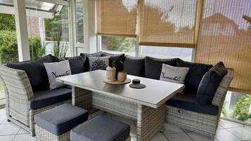 A room with a central table, cushioned seating, and large windows with blinds.