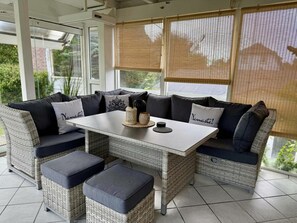 Una habitación con una mesa central, asientos acolchados y ventanas amplias con persianas.