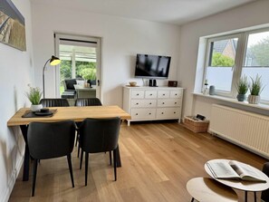 A modern living room with a wooden dining table, black chairs, a white cabinet, a flat-screen TV, and a window with plants. - Holiday Apartment With 1 Bedroom (Sylt)