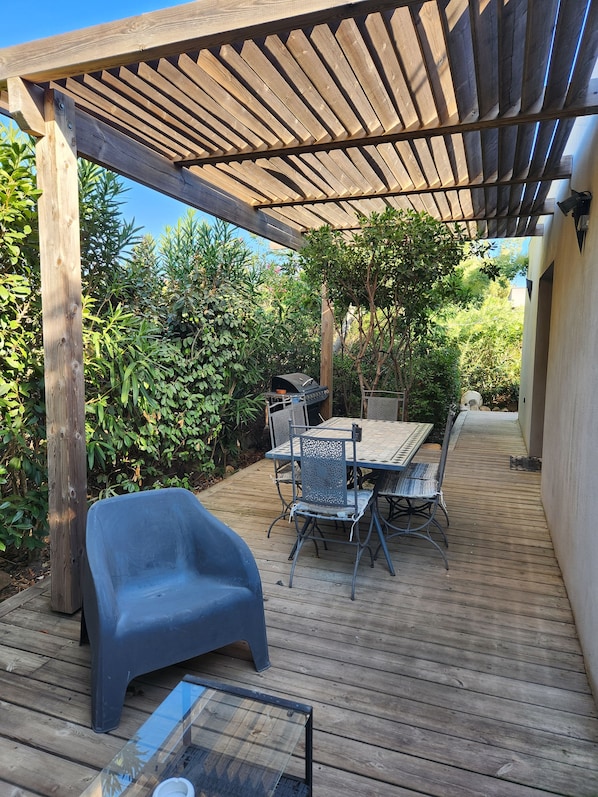 Une terrasse en bois avec une pergola, une table entourée de chaises et une chaise en plastique.
