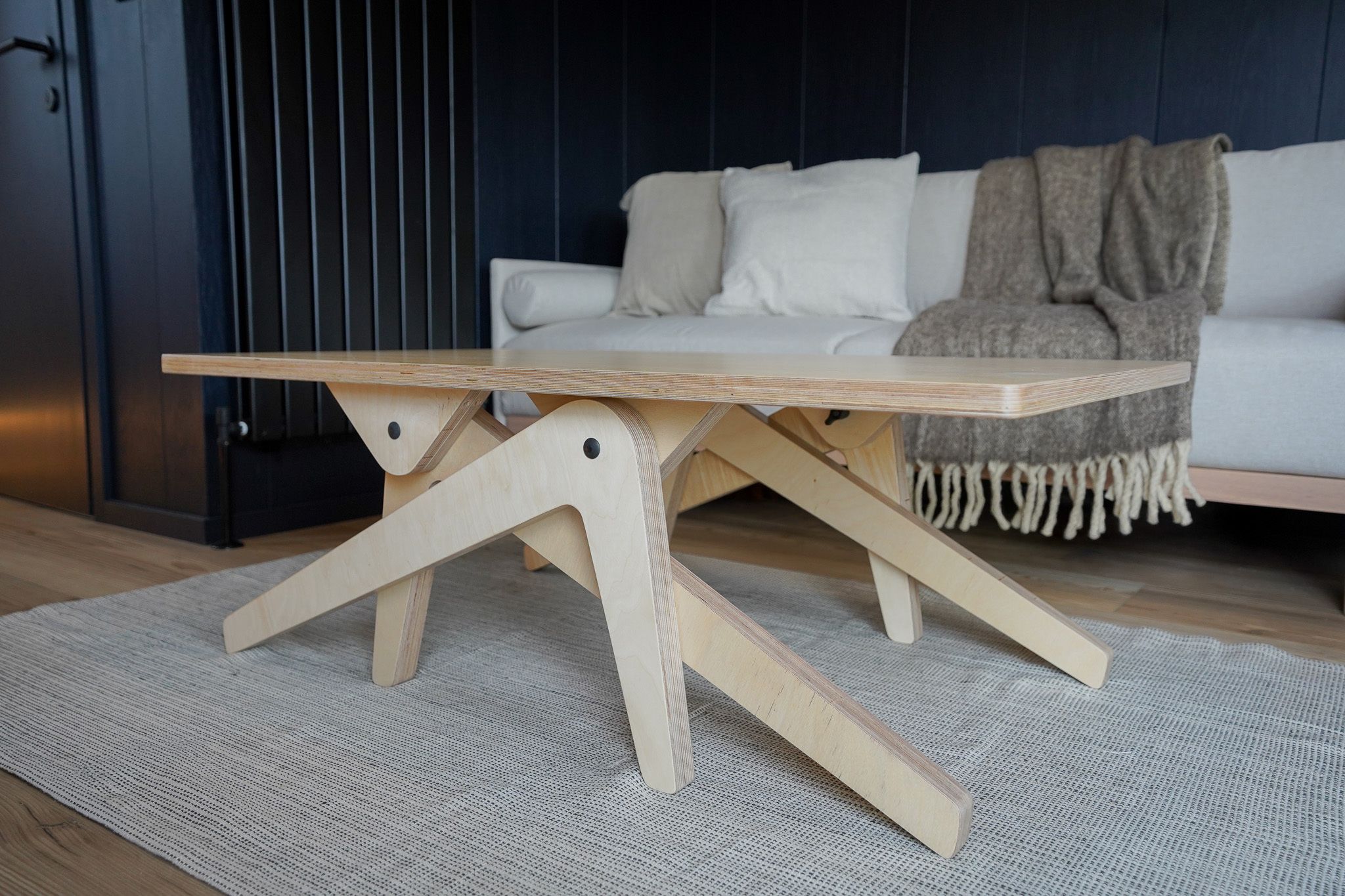 A wooden table with a unique X-shaped base design, placed on a textured rug in a room with a dark wall and a white sofa.