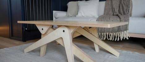 A wooden table with a unique X-shaped base design, placed on a textured rug in a room with a dark wall and a white sofa.