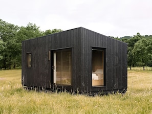 Eine kleine, dunkle Holzhütte mit einem Fenster und einer Tür, gelegen in einem grasbewachsen Feld mit Bäumen im Hintergrund.