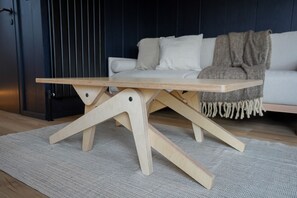 Une table en bois dotée d’un pied original en forme de X, posée sur un tapis texturé dans une pièce aux murs sombres et où se trouve un canapé blanc.