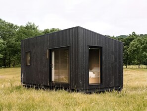 Eine kleine, dunkle Holzhütte mit einem Fenster und einer Tür, gelegen in einem grasbewachsen Feld mit Bäumen im Hintergrund.