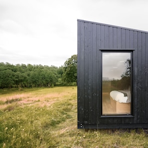 Une cabane moderne aux couleurs sombres, dotée d’une grande fenêtre, située dans un champ verdoyant avec des arbres en arrière-plan.