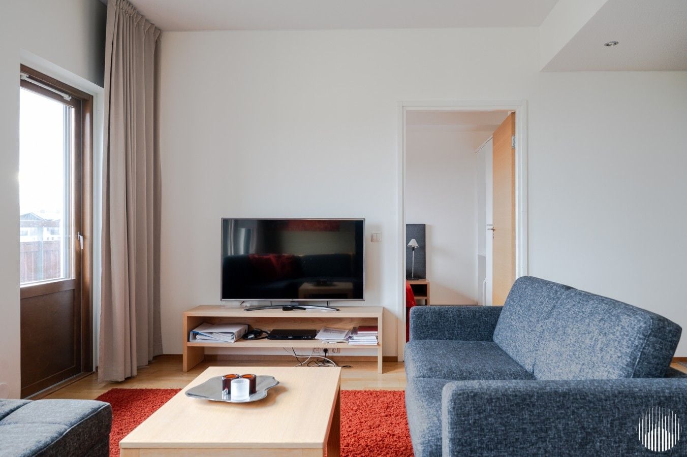 Un salon moderne avec un téléviseur à écran plat posé sur un meuble en bois, un canapé gris, un tapis rouge et une petite table avec un plateau.