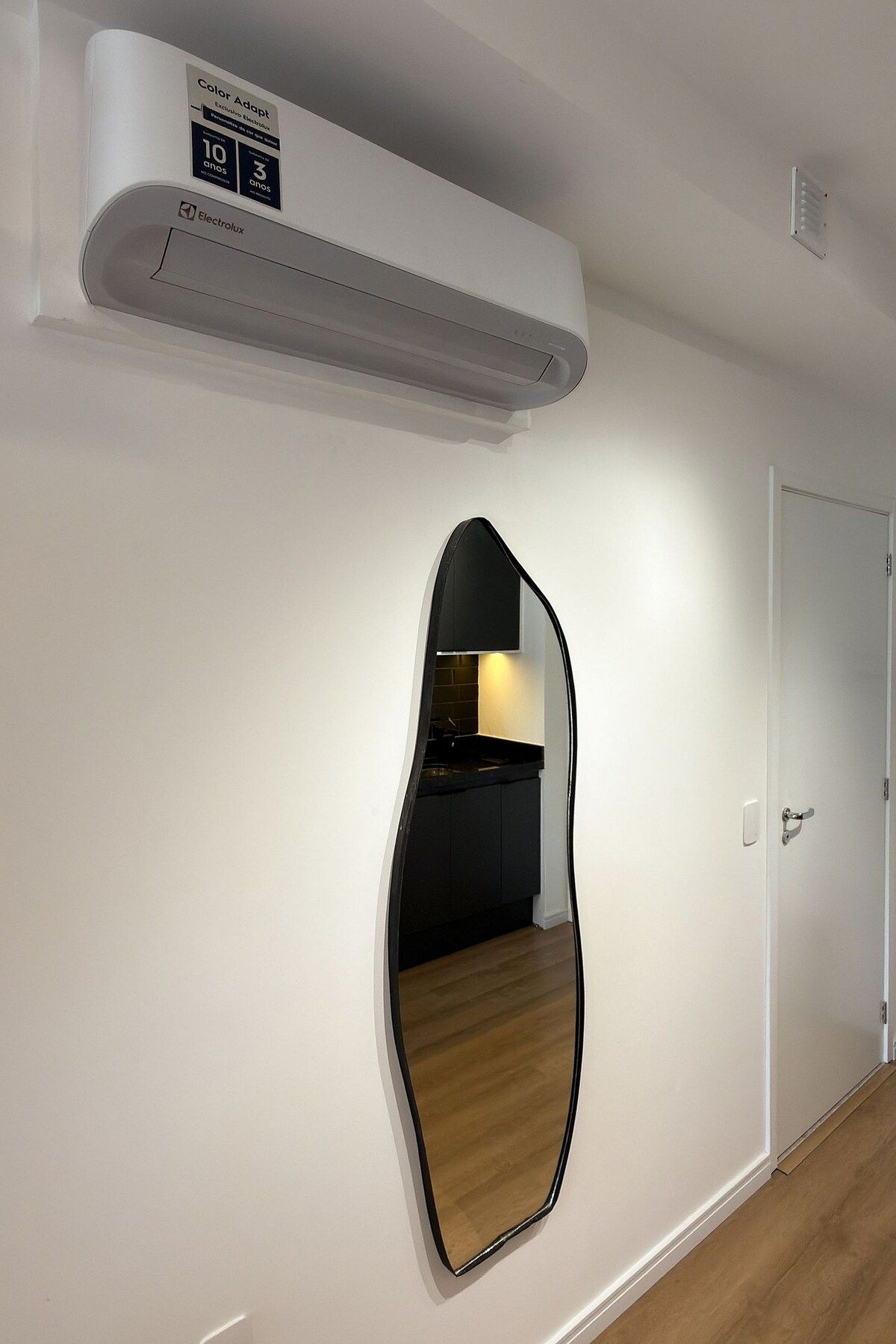 A hallway with a white wall, a black-framed mirror, and a white ceiling with an air conditioning unit.