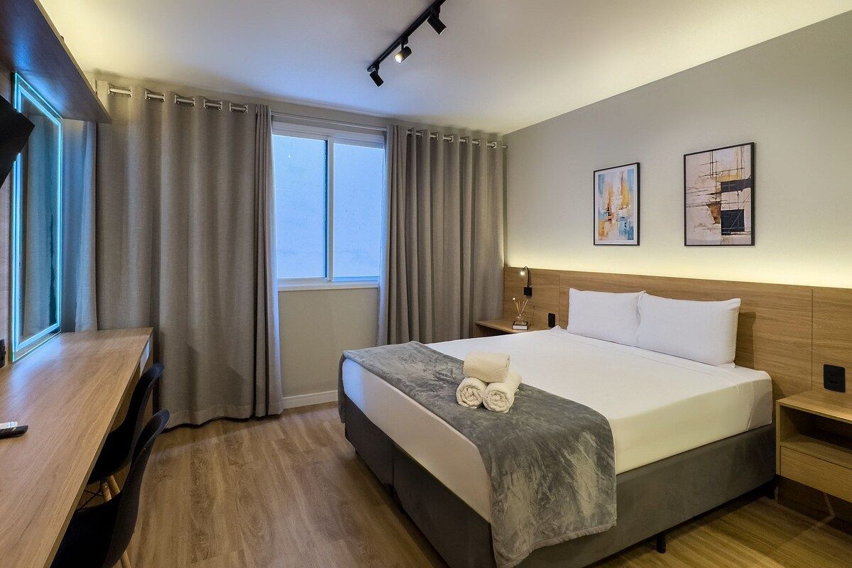 Een hotelkamer met een bed, een bureau, een stoel, een nachtkastje, een lamp en twee schilderijen aan de muur.