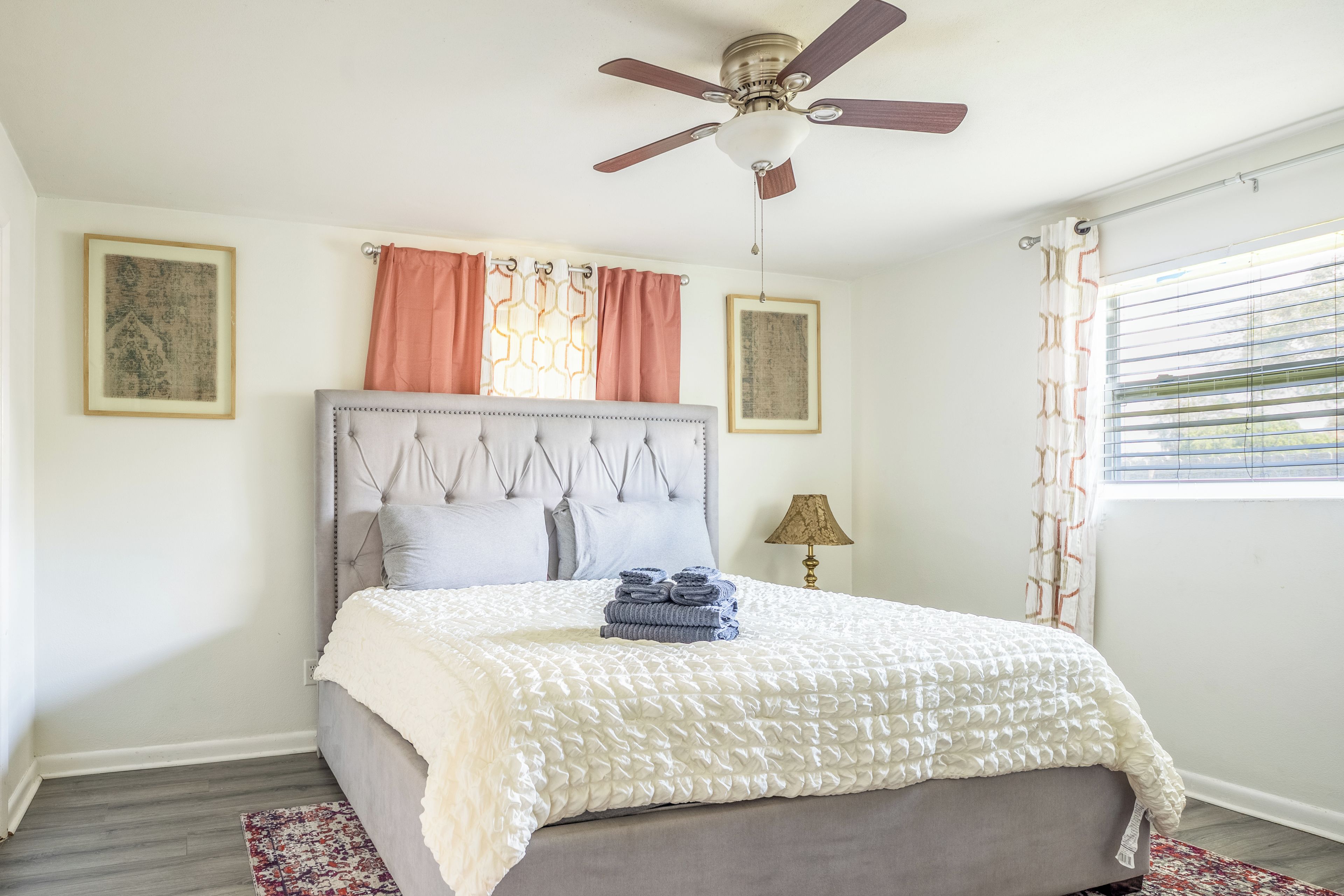 Quarto com cama, ventilador de teto, janela com persianas e duas molduras na parede.