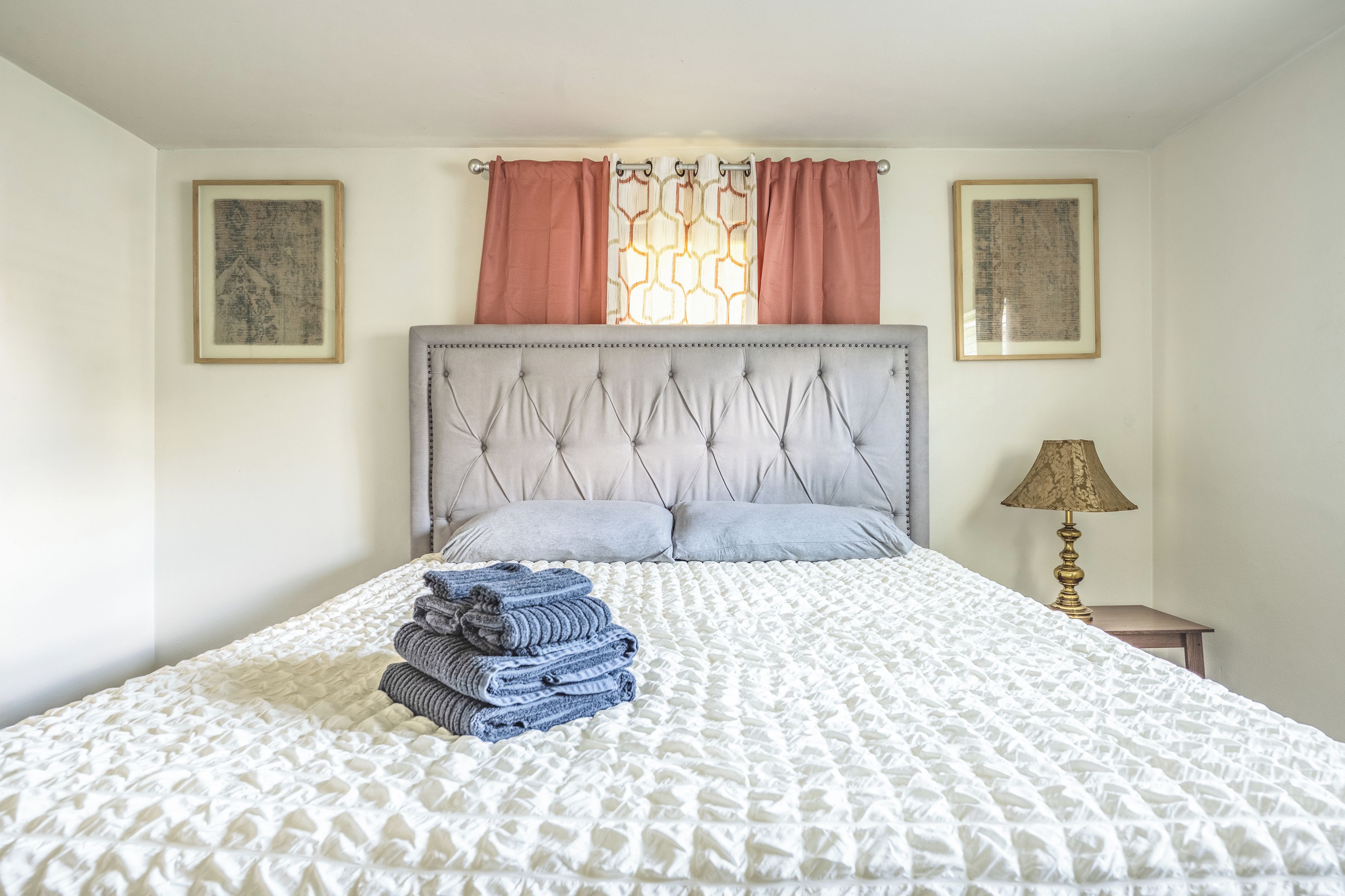 Una cama bien hecha con una colcha blanca y gris estampada, una pila de toallas dobladas y un cabecero con diseño capitonado.