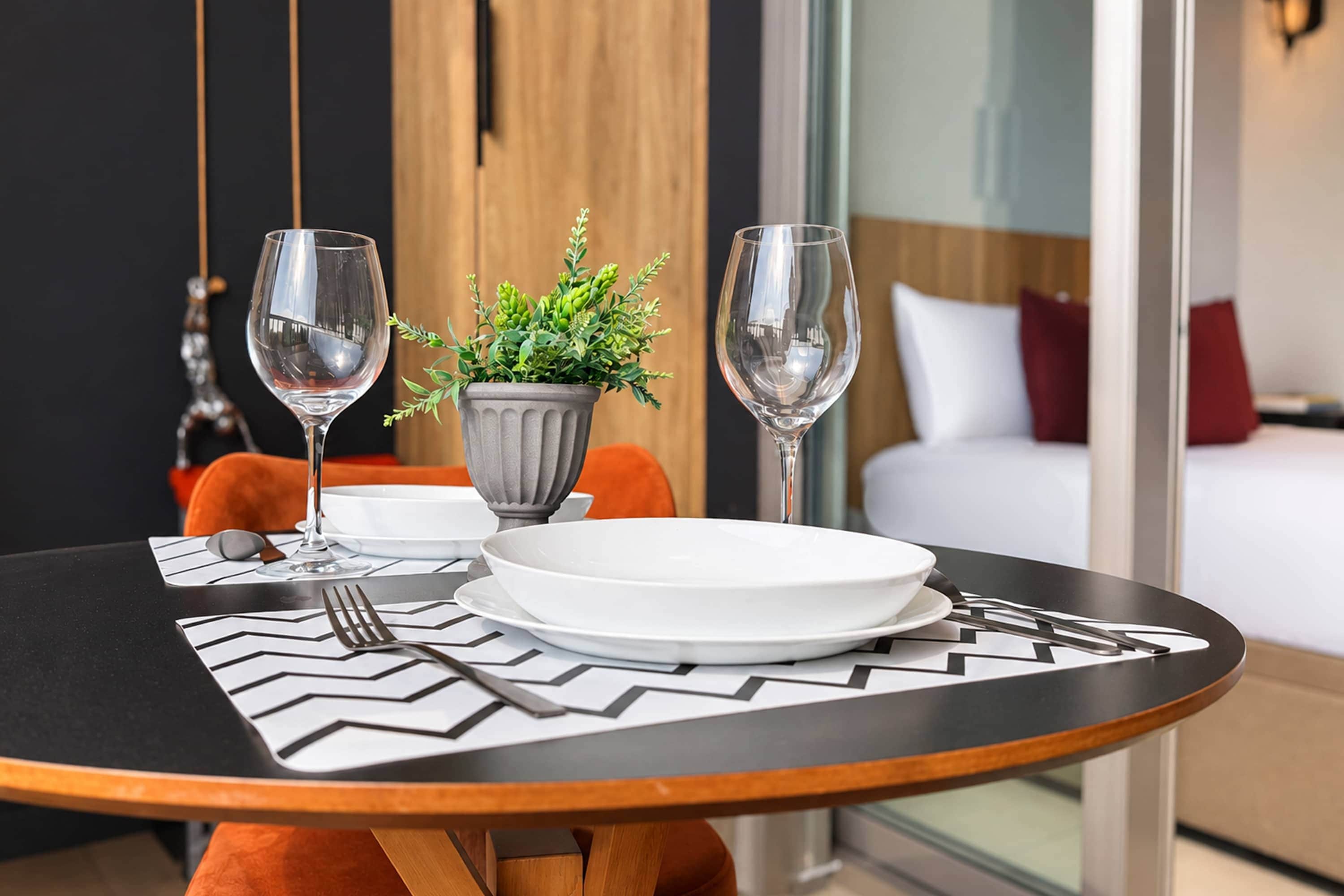 A hotel room with a dining table set for two, a potted plant, and a bed with a red pillow.