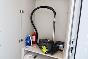 Huoneessa on pölynimuri, sammutin ja sininen kassi hyllyllä.