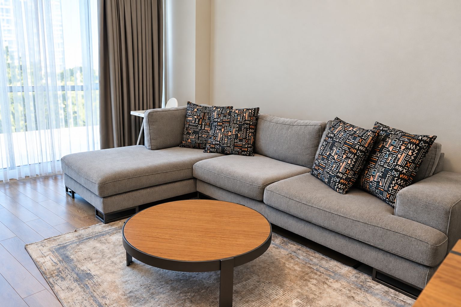 Una sala moderna con un sofá seccional gris, almohadones estampados, una mesa de centro redonda de madera y un ventanal con cortinas.