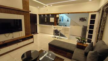 A modern living room with a flat-screen TV, a sofa, a coffee table, and a dining area with a TV and a small table.