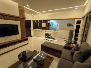 A modern living room with a flat-screen TV, a sofa, a coffee table, and a dining area with a TV and a small table. - Kaattil Homes - Luxury Villa & Apartments (Varkala)