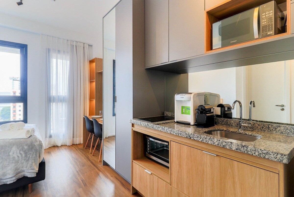 A compact hotel room with a kitchenette, a bed with a white duvet, a dining area with a table and chairs, and a large window with sheer curtains.