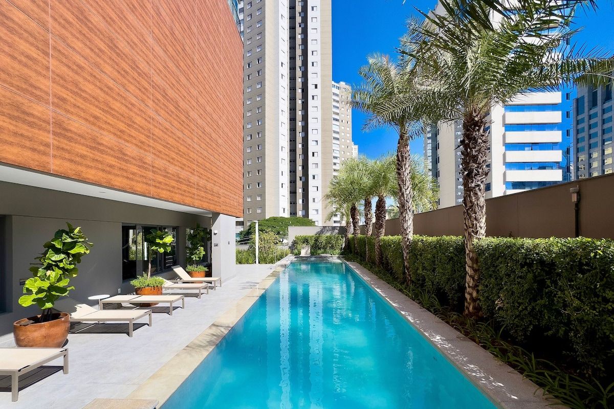 A swimming pool with lounge chairs and potted plants, surrounded by tall buildings and palm trees.