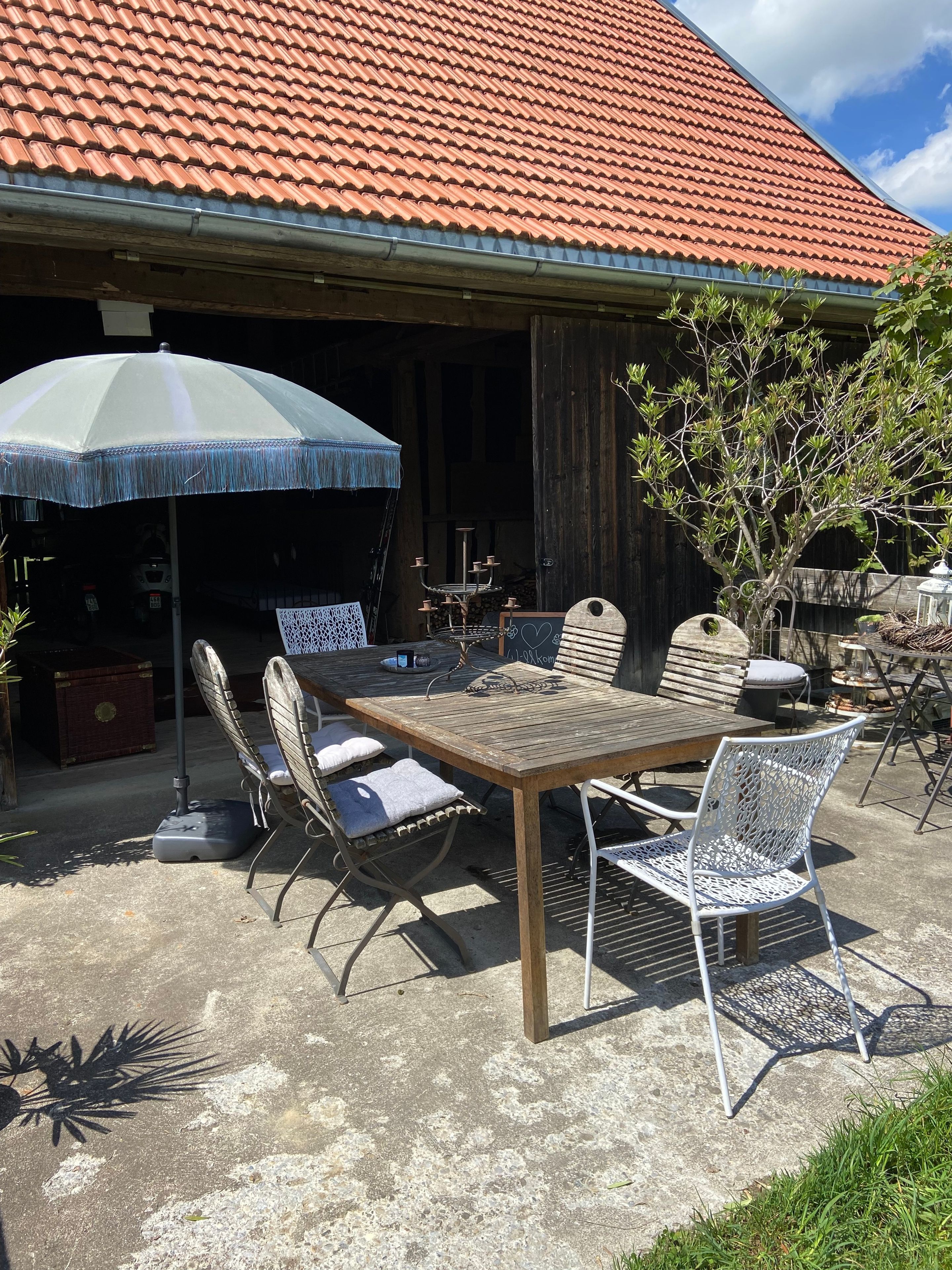 An outdoor patio with a wooden table and chairs, a metal umbrella, and a red-tiled roof.