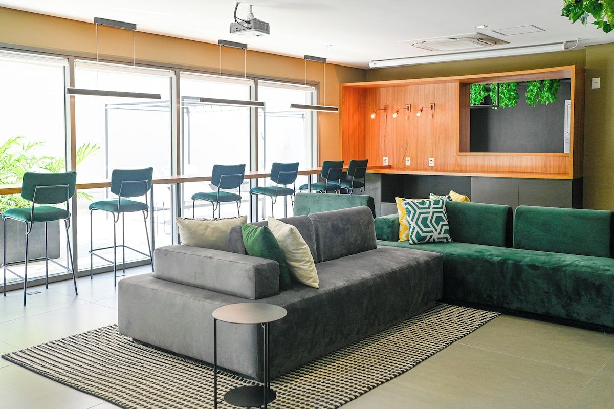 Un espace salon moderne avec un canapé d’angle gris, des fauteuils verts, une petite table ronde et un meuble en bois orné d’une plante verte.