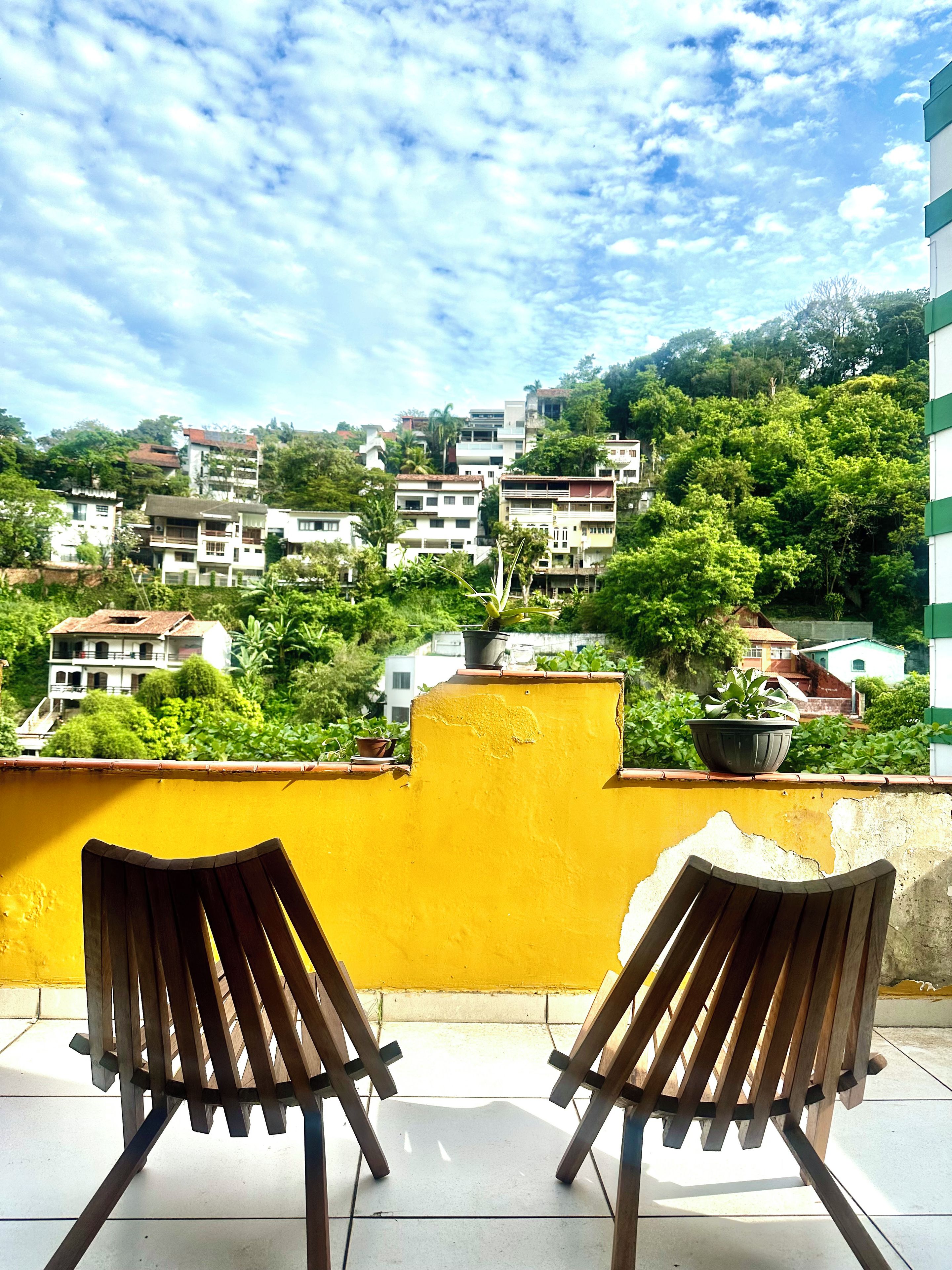 Dos sillas de madera en un balcón con una pared amarilla, con vistas a una colina con edificios y vegetación.