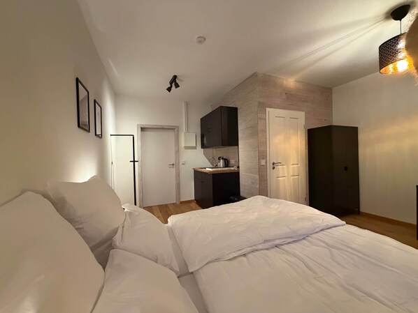 A bedroom with a bed, a kitchenette, and a door leading to another room.