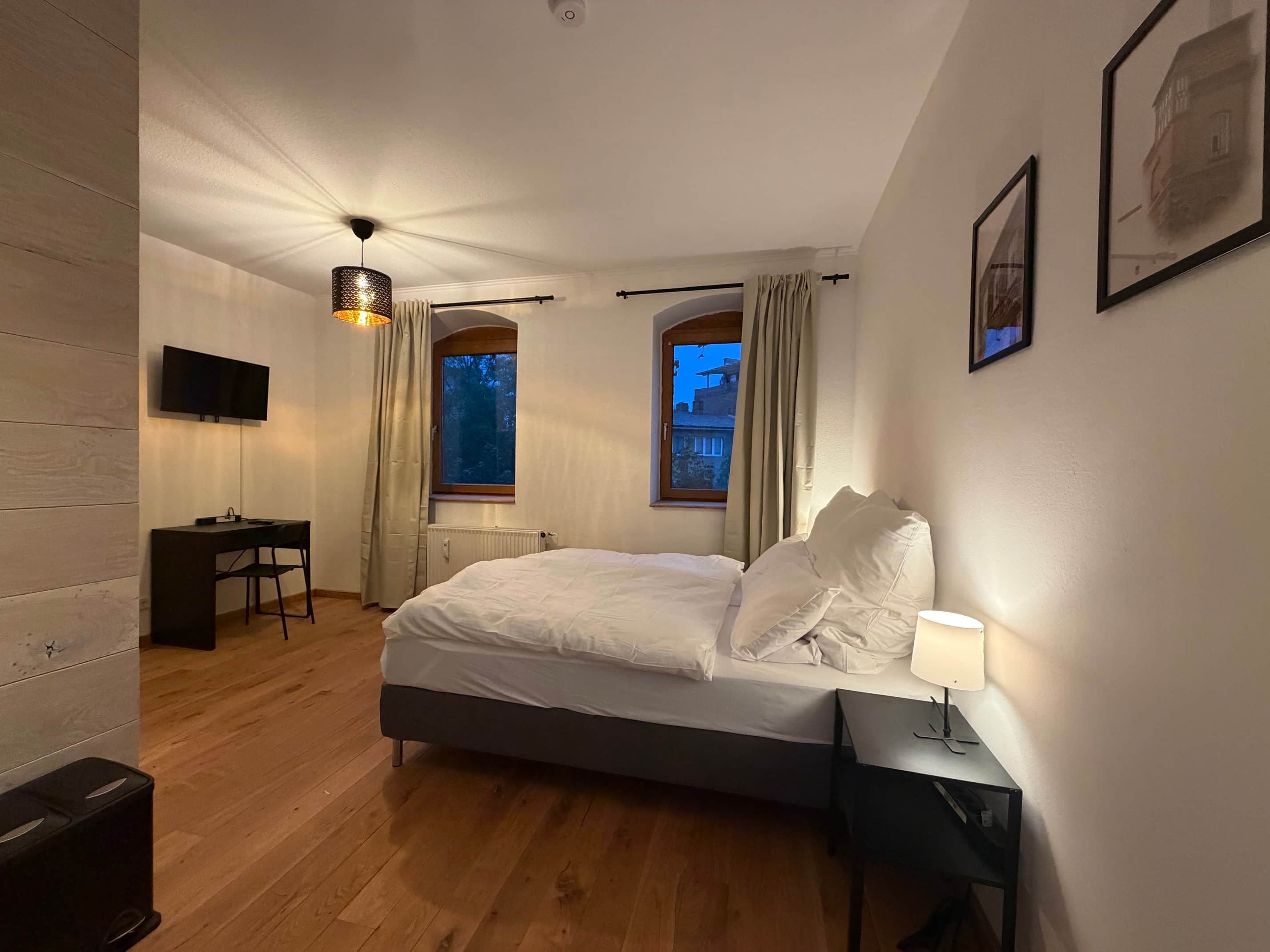 A bedroom with a bed, a desk, a chair, a lamp, and a television on the wall.