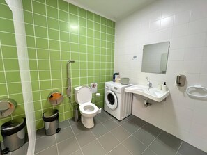 Kylpyhuoneessa on vihreät laatat, wc-istuin, pesukone, pesuallas ja roskakori.