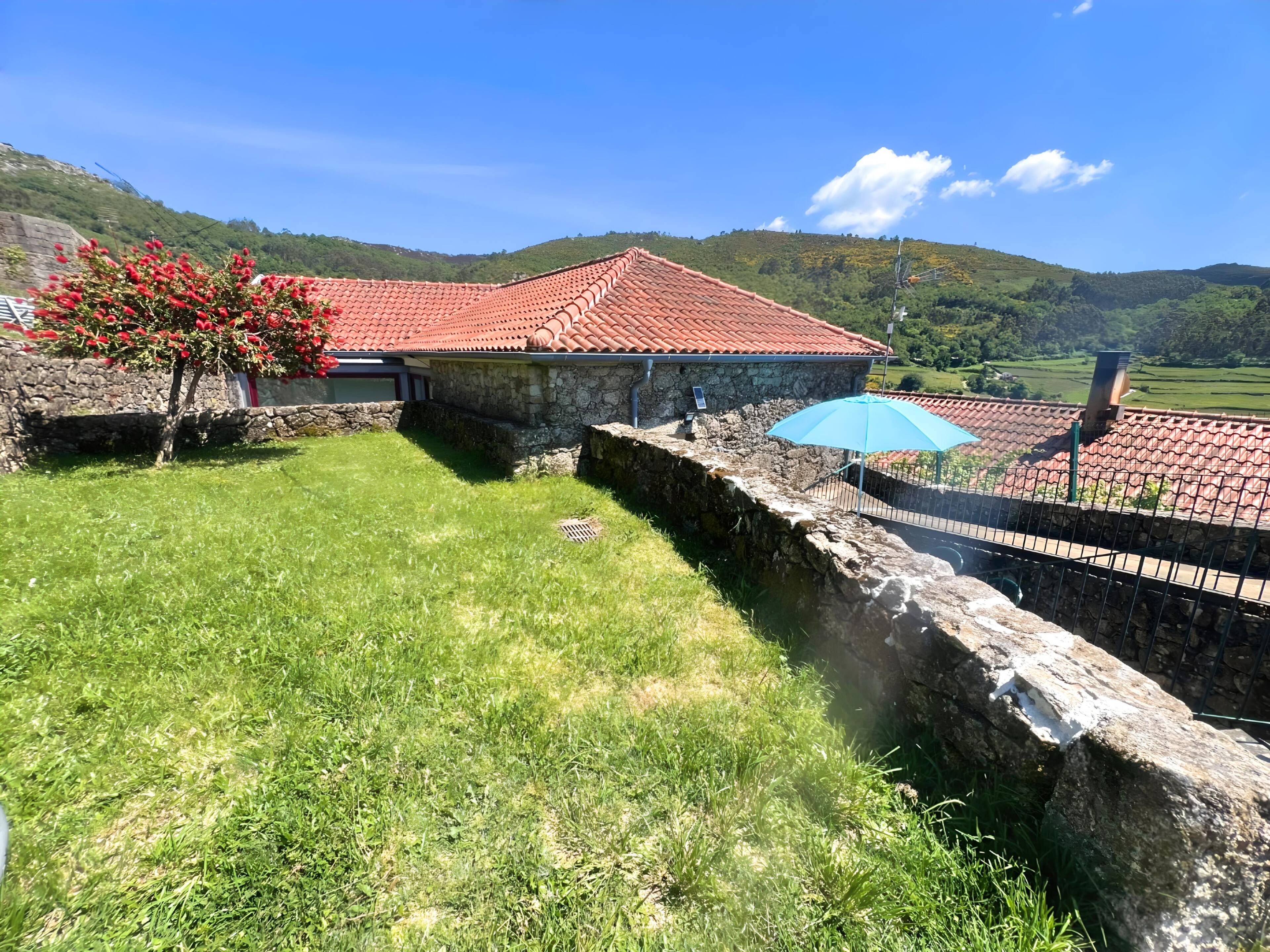 A stone building with a red-tiled roof, surrounded by a stone wall and a grassy area with a tree.