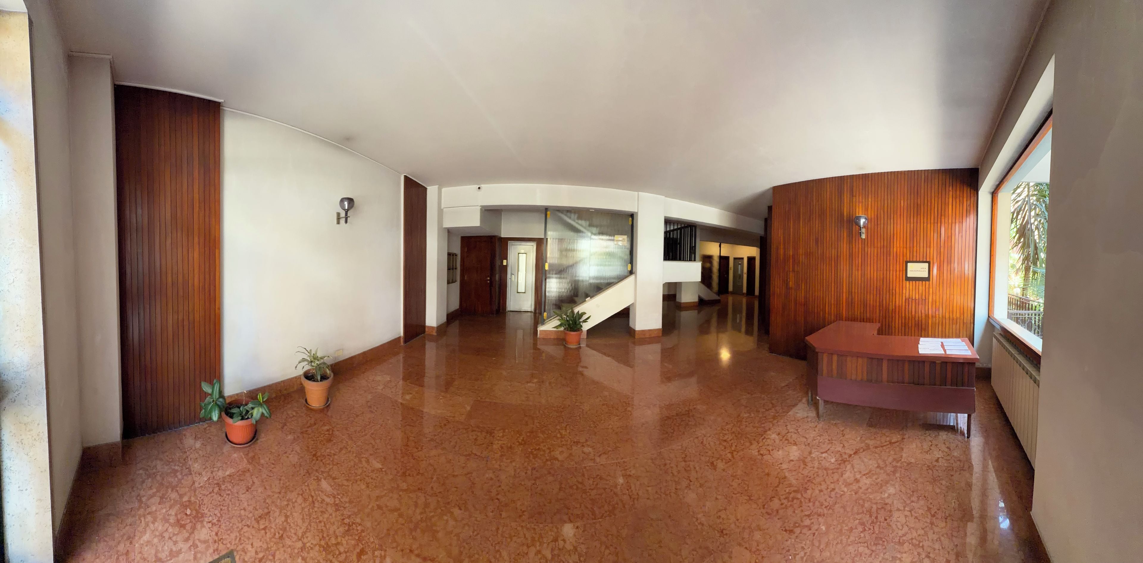 A spacious hallway with a polished floor, wooden paneling on the walls, a reception desk, and potted plants.