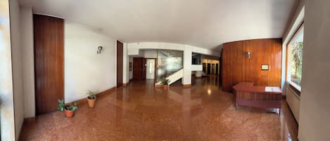 A spacious hallway with a polished floor, wooden paneling on the walls, a reception desk, and potted plants.