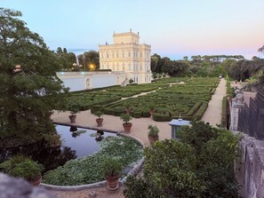 A formal garden with a symmetrical layout, a large building with classical architecture, and a body of water with lily pads. - New Listing (Roma)