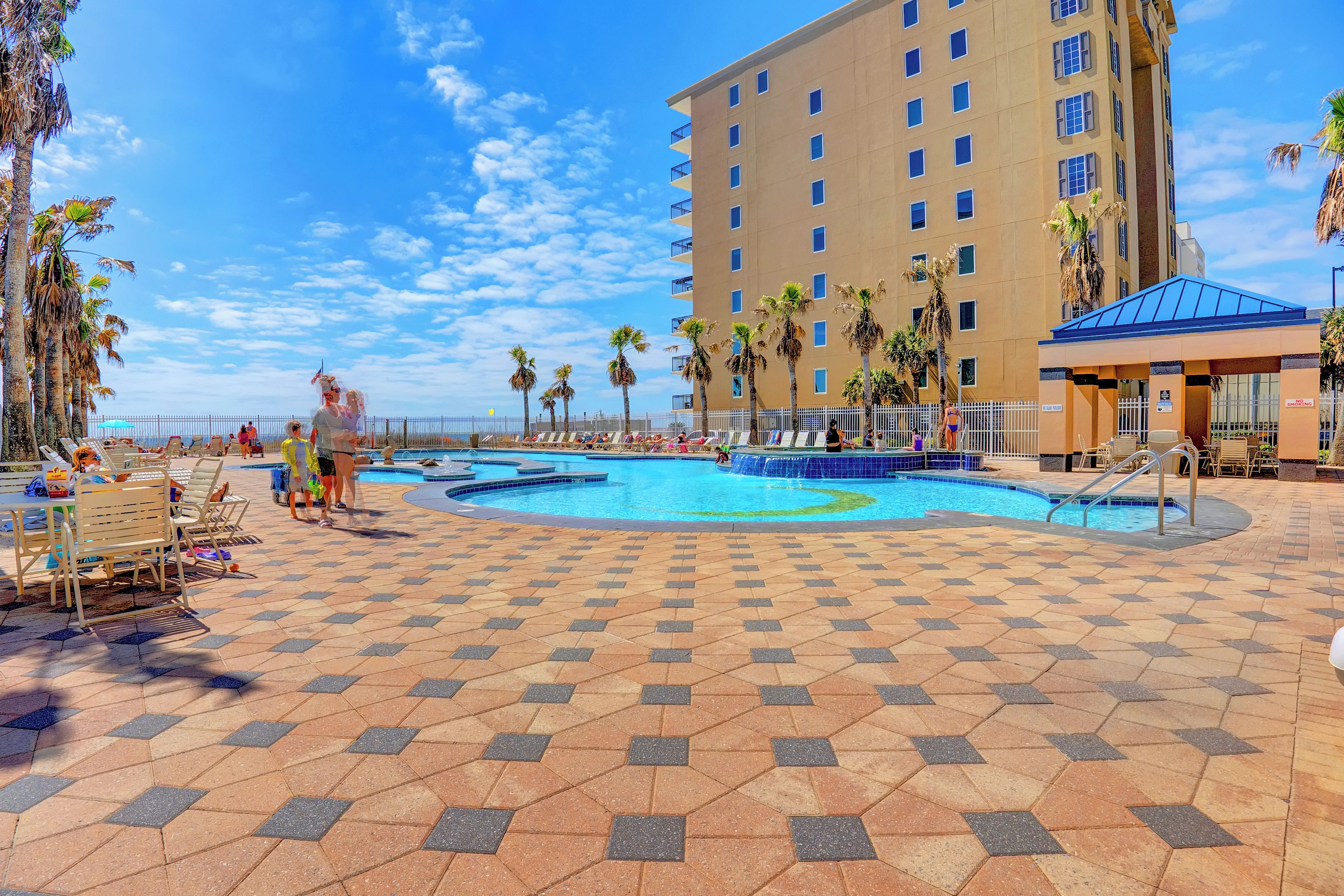 A pool area with tiled flooring, palm trees, and a tall building in the background.