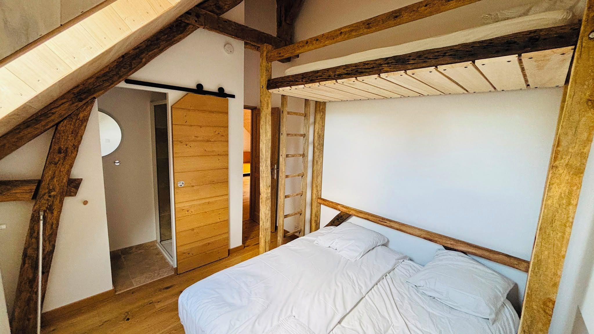 A bedroom with a sloped ceiling, wooden beams, a bed with white bedding, and a ladder leading to a lofted area.