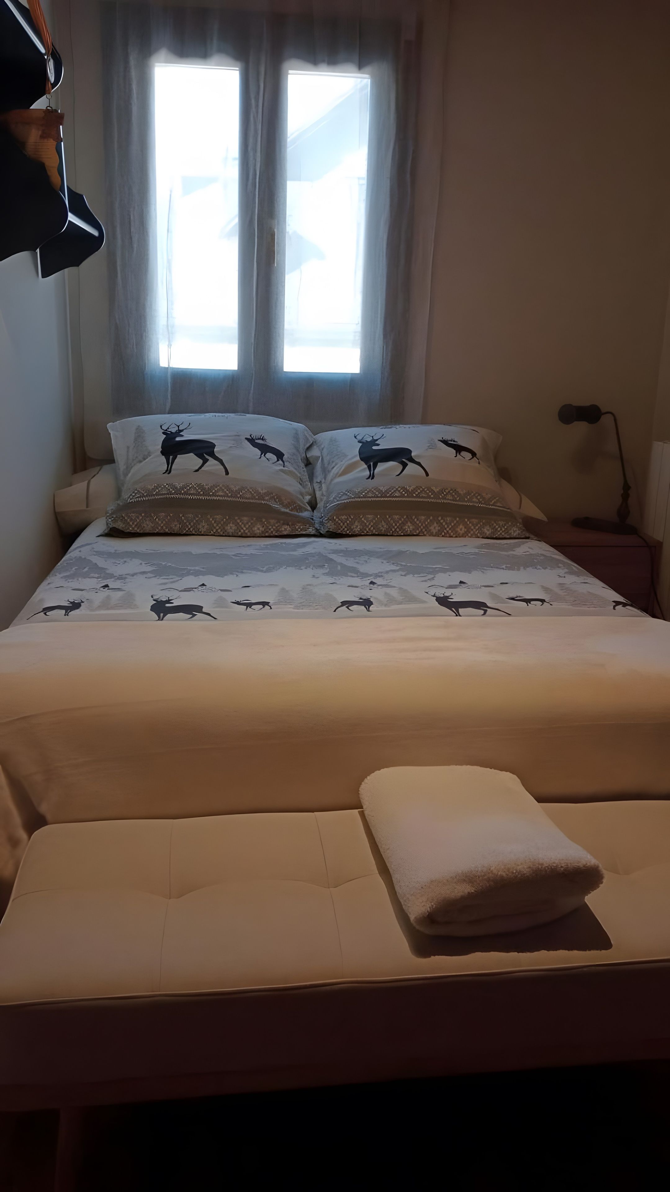 Een bed met een spreideken met hertenprint, een bedlamp en een raam met gordijnen.