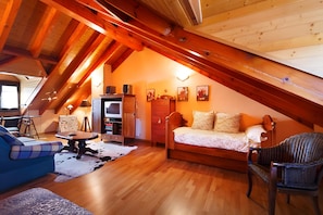 Ein gemütliches Dachzimmer mit Holzbalkendecke, einer Couch, einem Sessel, einem kleinen Tisch und einem Fernseher.