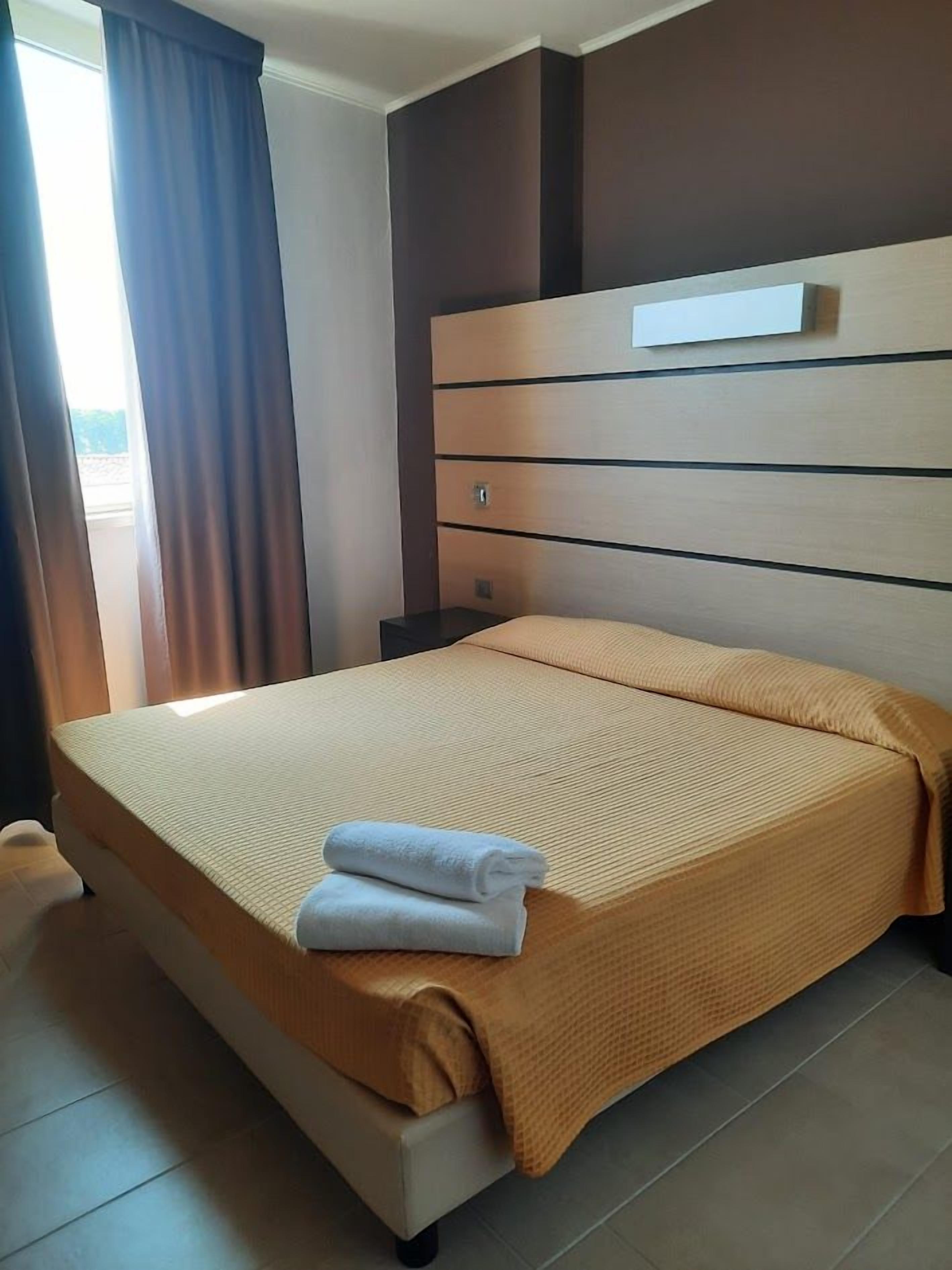 Une chambre d’hôtel avec un lit, une table de chevet, une étagère fixée au mur et une fenêtre avec des rideaux.