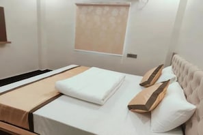 A neatly made bed with a white and beige bedspread, two pillows, and a window with a patterned blind. - FabHotel Sarnath Inn (Varanasi)