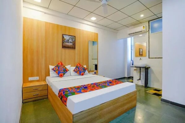 Habitación de hotel con cabecera de madera, cama con almohadas de colores, mesita de noche, espejo, ventana y baño con lavabo y espejo.