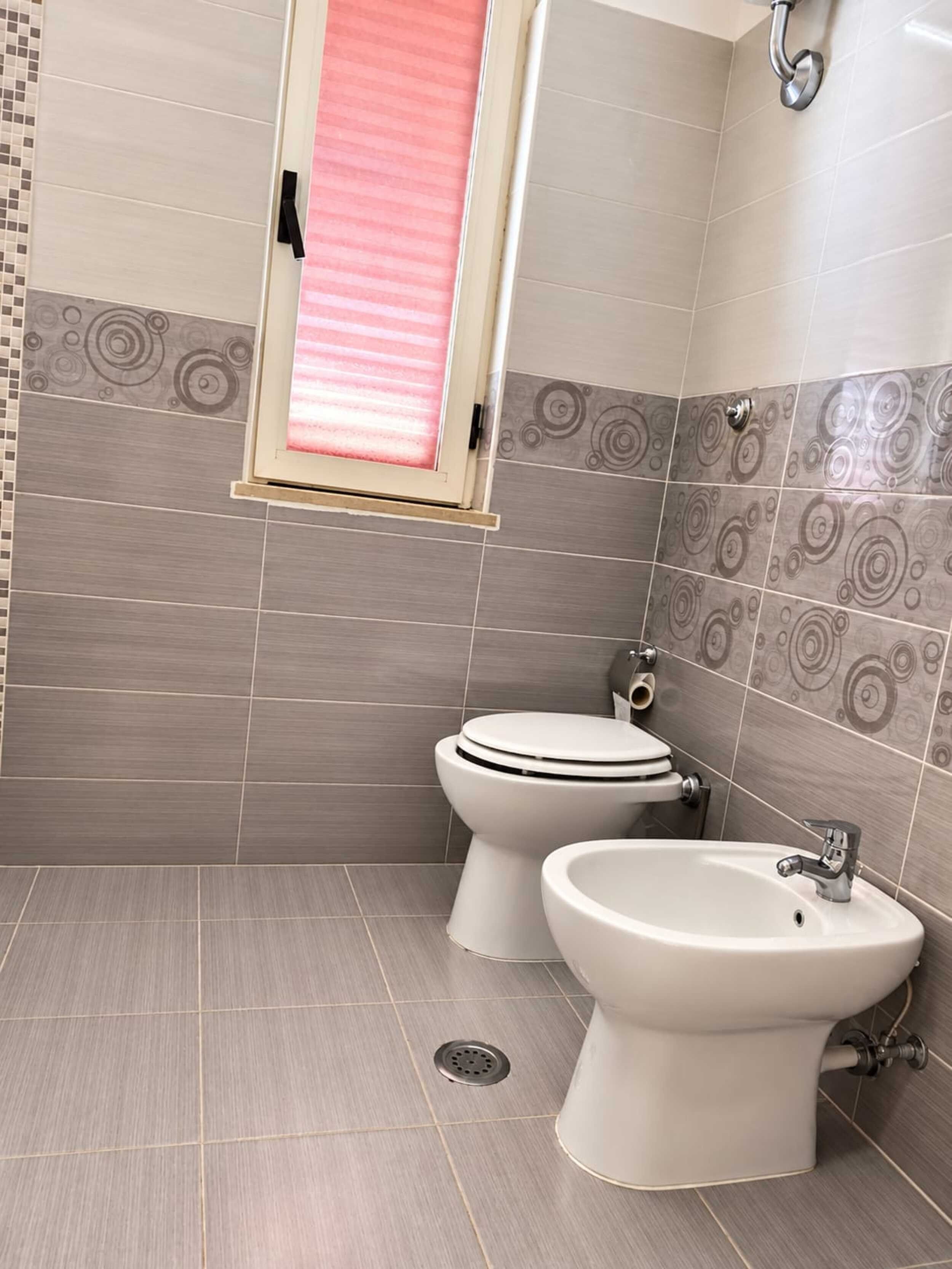 Une salle de bain avec deux toilettes, un bidet et une fenêtre avec un store rose.