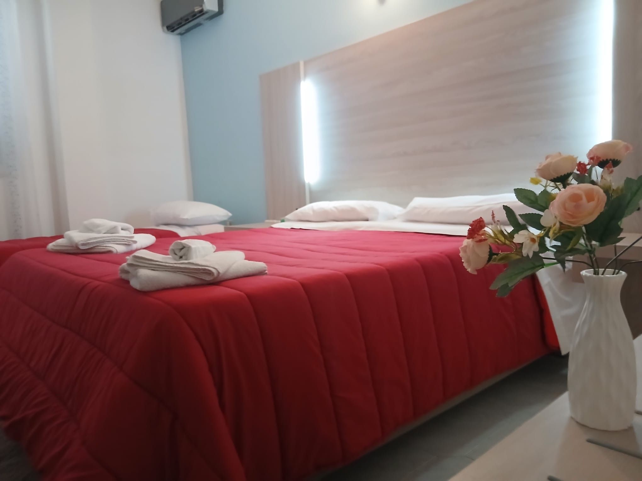 A hotel room with a bed, pillows, a vase of flowers, and a wall-mounted air conditioning unit.