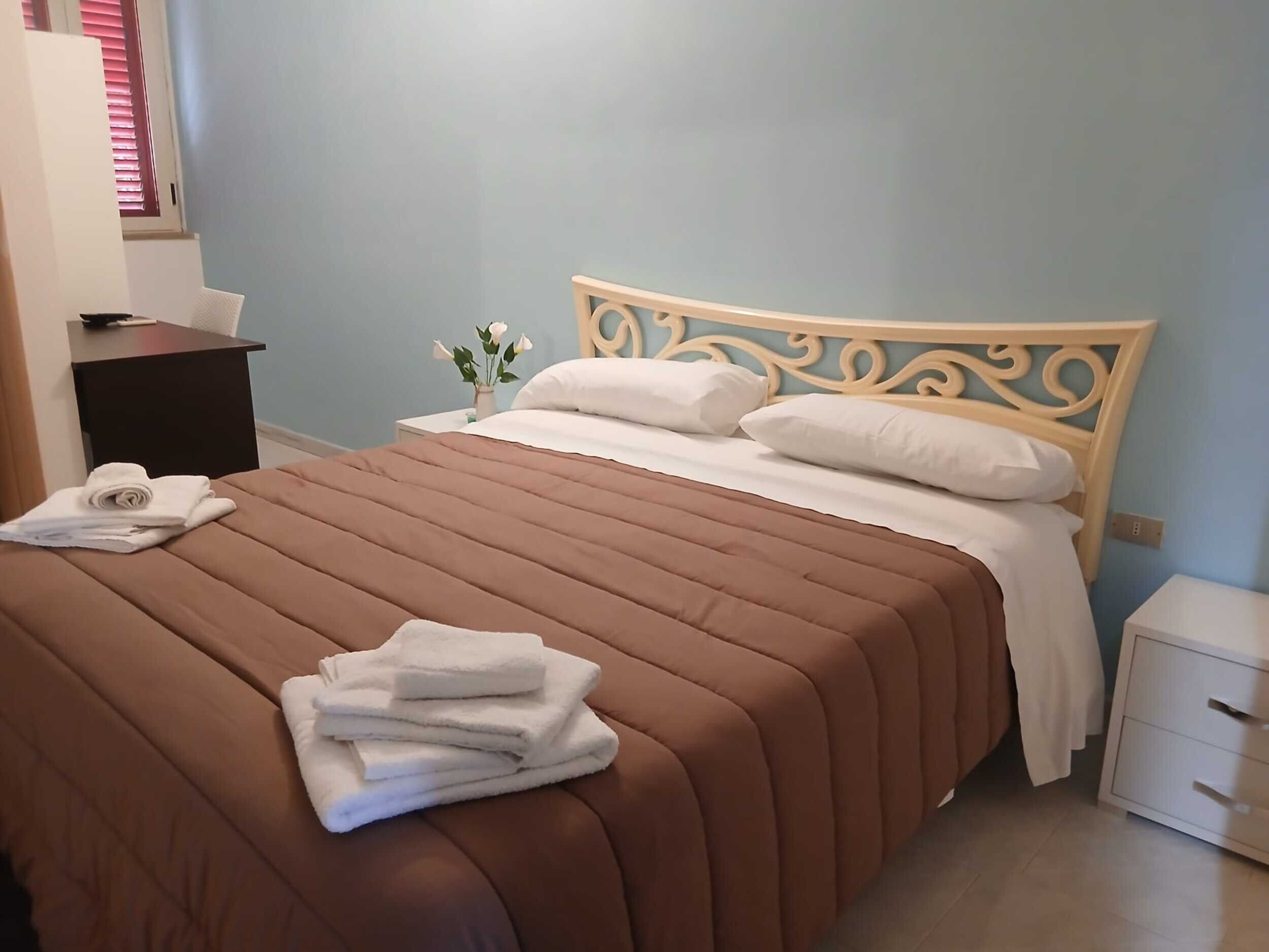 Ein ordentlich bezogenes Bett mit einer braunen Tagesdecke und weißen Kissen, ein kleiner Nachttisch mit einem Blumenstrauß und ein Beistelltisch mit einer Lampe.