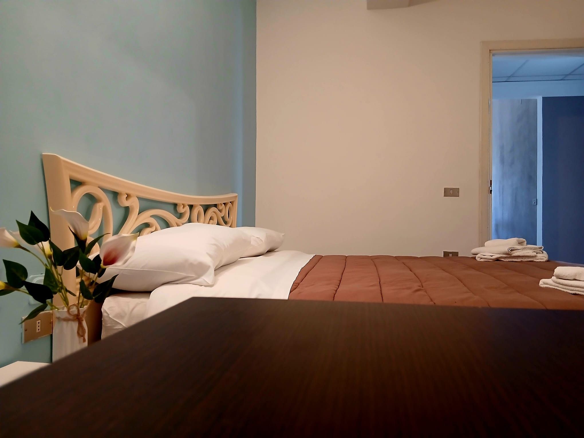 A bedroom with a wooden bed, white bedding, a vase with flowers, and a blue wall.