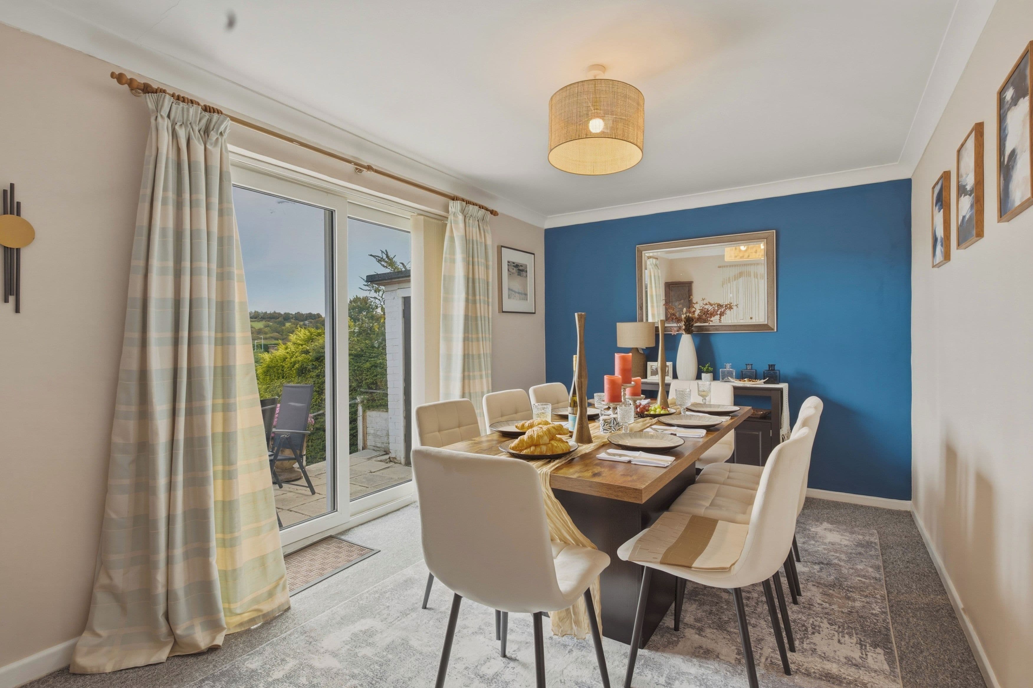 A dining area with a table set for four, chairs, a large window with curtains, and a ceiling light fixture.